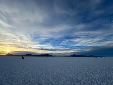 View of the vast Bonneville Salt Flats at sunset with scattered clouds in the sky and distant mountains on the horizon, featuring individuals walking and enjoying the serene landscape.
