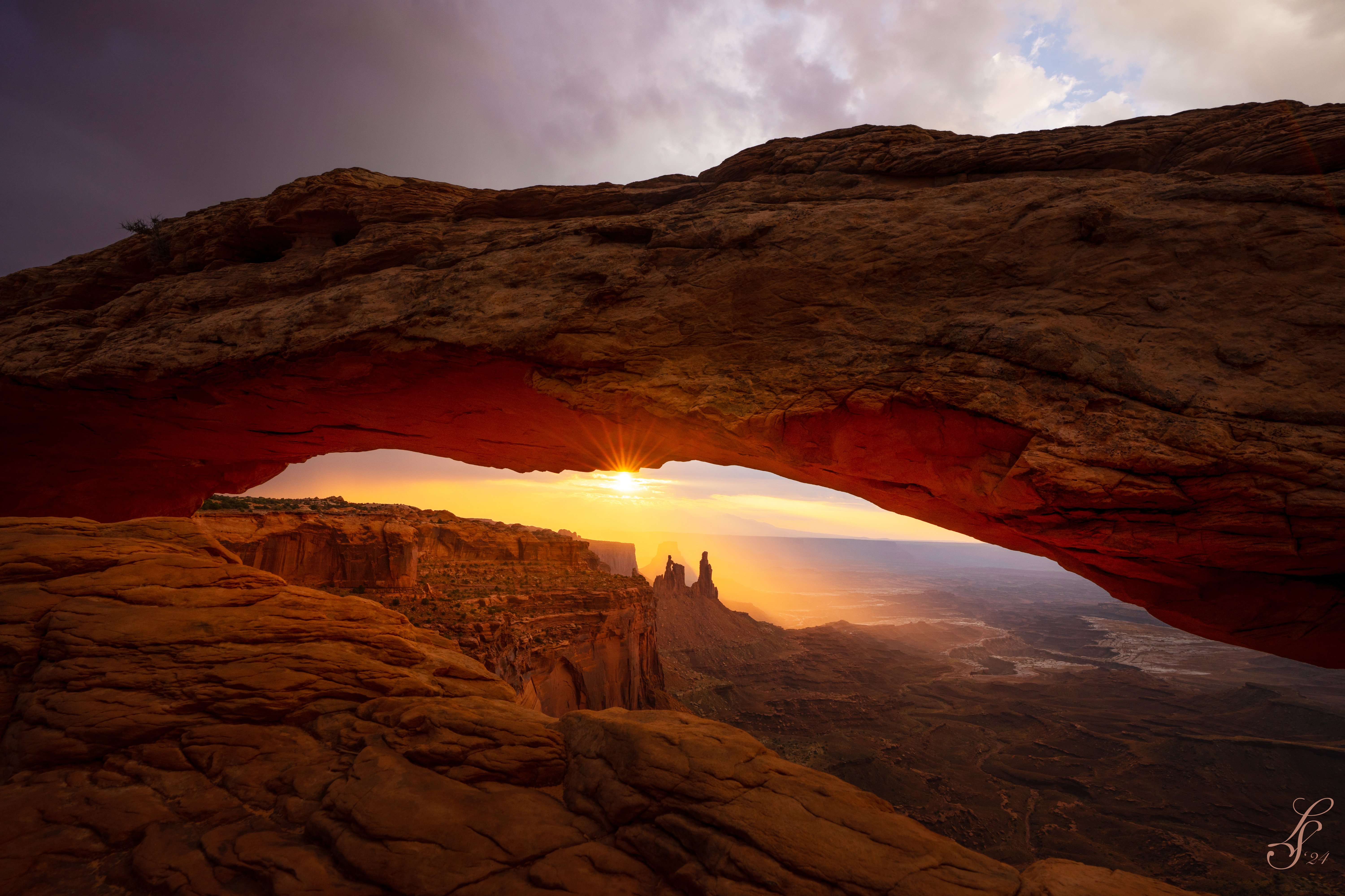 Sunrise view through Mesa Arch in Canyonlands National Park, showcasing warm light illuminating the underside of the arch with a canyon landscape in the background.