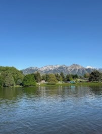 View of a l ake with the Wasatch mountaion range in the background, featuring clear blue skies and lush greenery along the shoreline.
