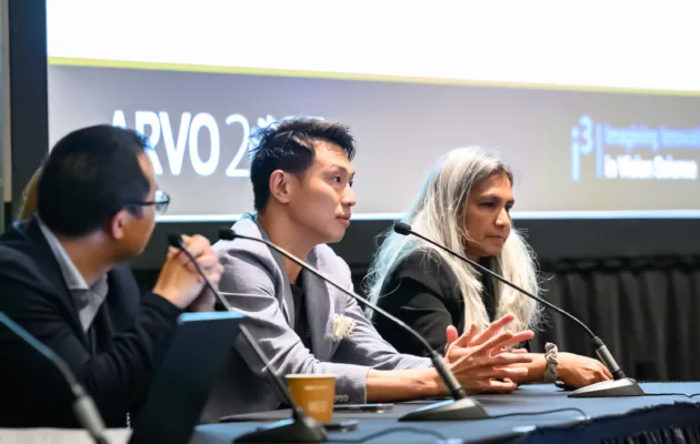 Three panelists seated on stage at a table with microphones and a large screen in the background
